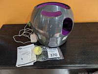 Silver and purple PetSafe Automatic Ball Launcher with power cable, yellow tennis ball, and user manual on wooden surface against orange background.