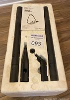 Top-down image of multiple accessories fitting in styrofoam, including two long black tubes, two nozzle attachments, and owner's manual.