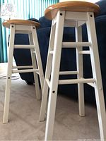 Close-up front view of two stools showing natural wood seats and white legs.