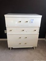 White vintage wooden dresser with four drawers, brass knobs, standing on angled legs against a wall. Shows signs of scratches and wear especially on the middle drawer which has a diagonal wood pattern.