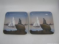 Photo of two matching square porcelain dishes featuring a mermaid sitting on a rock and a sailboat in the background, placed side by side.