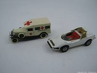 Photo showing side angle of two vintage die cast toy cars - cream colored Lledo ambulance with medical red crosses and white Corgi Alfa Romeo P33 toy car with red interior and gold roll bar.