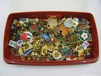 Wide shot of approximately 60 assorted old pins inside a reddish-brown Tupperware container, showing a variety of colors and styles of lapel/pins.