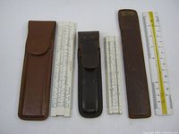 Three old pocket rulers laid out with their respective leather cases, showing scale and wear.