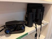 Two pairs of binoculars placed on a white shelf: one pair in a black zippered case and the other standing beside it with a carrying strap.
