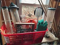 Red plastic basket with multiple clear plastic tube bird feeders with green caps and perches. Green rubber gloves visible inside basket, wire cage feeder also present.