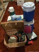 Overview of camping supplies showing coolers, water dispenser, thermos, picnic basket with blankets, ice packs, folders, and small bag.