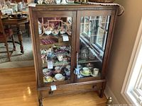 Full front view of wooden china cabinet with glass doors and bottom drawer. Cabinet is filled with various china pieces (not included in lot).