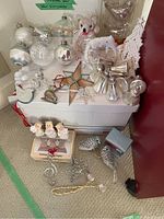 Photo showing a variety of vintage Christmas ornaments including glass bells, glass birds, stuffed bear, star sun catcher, singing angels in box, and a white moose figure, along with some textile and light decorations.