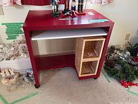 Front view of red computer desk showing the sliding white keyboard tray fully pushed in, with two woven basket storage cubes on the right side under the desk and caster wheels visible.