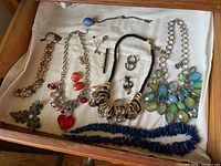 Wide view of drawer with costume jewelry: necklaces, earrings, pins including a blue beaded necklace, multi-colored stone necklace, black cord bead necklace, coral bead necklace and multiple pins and earrings.
