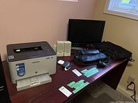 Samsung Xpress C410W printer, Toshiba monitor, Logitech and Soundworks speakers, Bang & Olufsen power bank, Apple wired headphones and black computer bag on a wooden desk near a window