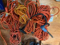 Several coiled orange and yellow outdoor extension cords of various lengths and plug types on wooden floor.