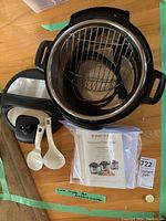 Top view of Instant Pot showing inside with metal cooking rack and black lid beside it, two white plastic spoons and a plastic bag with manual and recipes.