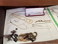 Overview of various necklaces, bracelets, earrings on white cloth, including pearl necklace, crystal necklace, brown/beaded necklace, small chains, earrings, and jewelry pieces