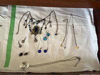 Overview of three necklaces, three pairs of earrings arranged on white cloth including large silver-tone necklace with blue beads and charms, various delicate chain necklaces, and sets of earrings