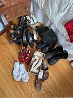 Overall view of 11 pairs of women's shoes on hardwood floor including black, beige, red shoes, black boots, white slippers, and brown sandals.