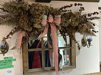 Front view of wood framed mirror with faux dried flower arrangement and pink ribbon bow, and two brass wall candle sconces with blue glass holders mounted on either side.