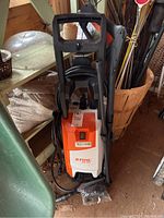 Front view of Stihl RE 90 pressure washer showing orange and white body, high-pressure hose coiled, spray gun mounted on side.