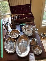 Photo showing assorted silver-plated cutlery in a wooden case, two oval serving trays, sugar shaker, carving set, candelabra, and small bowls on a table by a window.