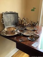 Overview of multiple silver plated serving ware items including trays, teapots, and serving utensils arranged on a table against a plain wall.