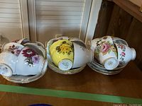 Photo showing 12 teacups and saucers arranged on a surface. Several different floral patterns including yellow, pink, and purple flowers.