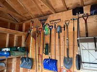 Wooden wall with various garden tools including shovels, rakes, pitchforks, pruning saw, tarps, and the wheelbarrow to the right