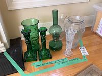 Photo showing four glass vases, one decorative green glass oil lamp, and two green glass candle holders arranged on the floor with handwritten labels.