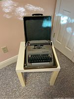Smith-Corona Clipper typewriter inside open carrying case on white side table next to wall and door.
