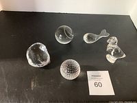 Photo showing five clear crystal decorative pieces arranged on a black surface with auction lot number 60: Wedgwood whale, Baccarat bird, Tiffany crystal tennis ball, Galway potato, Waterford tennis ball.