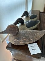 Three wooden duck decoys placed on a dark wooden surface against a white curtain background. The front duck shows significant paint wear and rough texture, while the two behind it retain more distinct black and gray paint patterns.