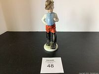 Front view of Tom Thumb Hungarian figurine showing a boy in feathered hat, blue vest, red pants, black boots on white base.