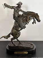 Front angled view of the bronze statue showing cowboy on bucking horse with plaque reading 'Wooly Chaps by Frederic Remington'