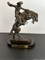 Front view of bronze sculpture showing cowboy on bucking horse with wool chaps, engraved base plaque visible