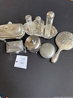 Photo showing the entire lot including silver butter dish, two trinket boxes, three glass bottles with silver lids on a tray, round compact, and hand mirror.