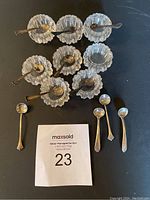 Photo showing 8 crystal salt dishes arranged in two rows with 11 sterling silver salt spoons placed on and near the dishes, some tarnishing visible on spoons.