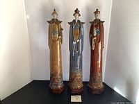 Front view of three tall ceramic Asian figurines in yellow, blue, and reddish-brown colors showing front details.