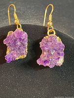 Pair of raw amethyst stone earrings mounted on gold tone backing with gold tone hooks, showing the front side with visible crystals and color variations in purple shades.