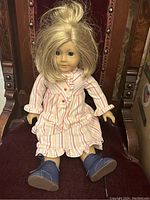 Photo of American Girl doll showing full frontal view seated on a dark red chair, dressed in striped dress and blue boots.