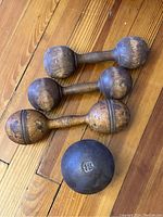 Photo showing three antique wooden dumbbells with varying levels of wear and one heavy iron shot put ball marked 12 pounds, all on a wooden floor.