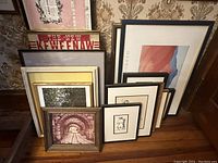 Collection of 11 variously sized framed artworks stacked against wall, diverse styles including sketches, prints, and text images.