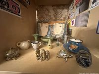 Wide view of all assorted miniatures and art tiles on display shelf, showing two art tiles of faces, small ceramic pitchers, and various figurines.