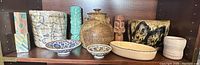 Wide view of shelf showing two pots, one container with lid, two small dishes with patterns, tiki-style pottery figurines, tall hexagonal vase with pastel colors and a small cream-colored pottery piece.