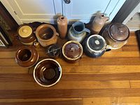 Overview of all bean pots and pottery bottles laid on hardwood floor in front of cupboard, showing shapes, lids, and colors.