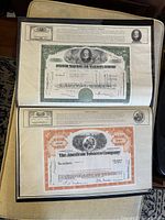 Top two pages of portfolio showing stock certificates of American Telephone and Telegraph Company (green border) and The American Tobacco Company (orange border). Each certificate has detailed historical context at the top.
