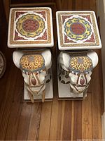 Pair of white decorative elephant stands with flat tops and colorful ornamental designs viewed from above showing tops and heads.