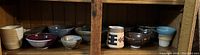 Wide view of multiple assorted pottery bowls, cups, and vessels arranged on a wooden shelf.