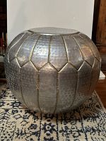 Side angle view of round metal pouf showing textured surface and vertical seams with gold tone accents.