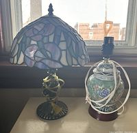 Photo showing two small lamps placed on a surface by a window. Left lamp has a stained glass Tiffany-style shade and metal base, right lamp is ceramic with floral motifs and wooden base. Both have cords attached.