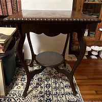 Front and side angled view showing carved edges, curved legs and circular lower shelf of the wooden table in a room.